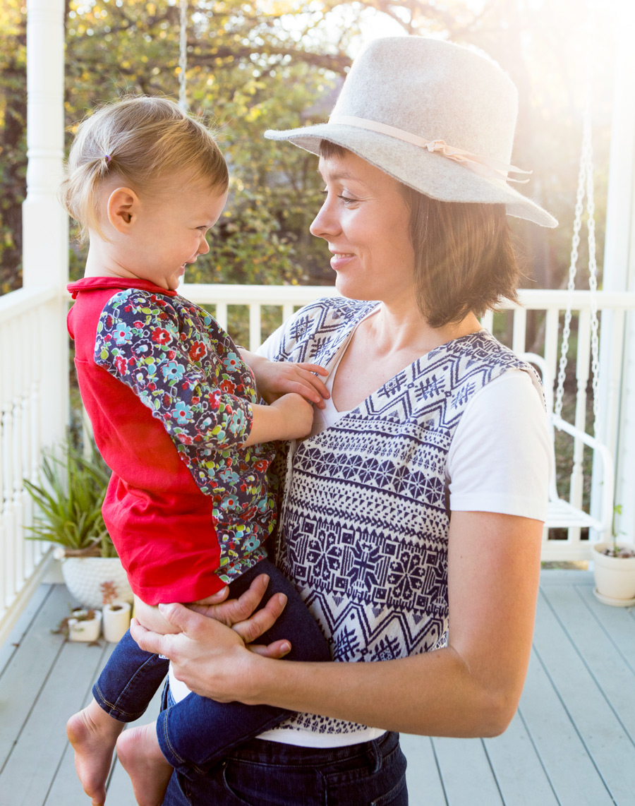 Mom and young daughter laughing on porch with sun steaming in and hat
